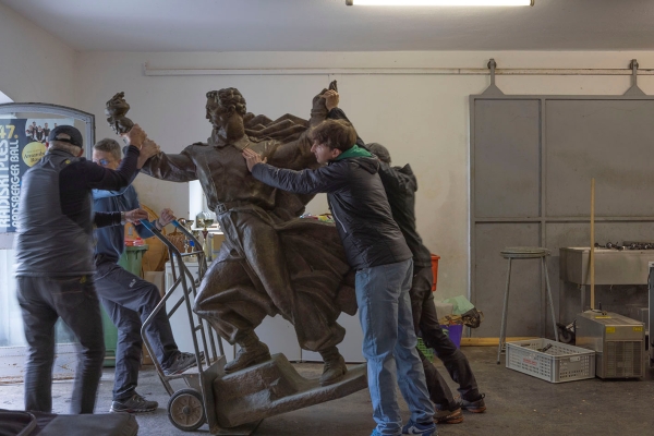Ein Denkmal einer männlichen Figur wird von einer Gruppe Männer mit Sackkarre transportiert.