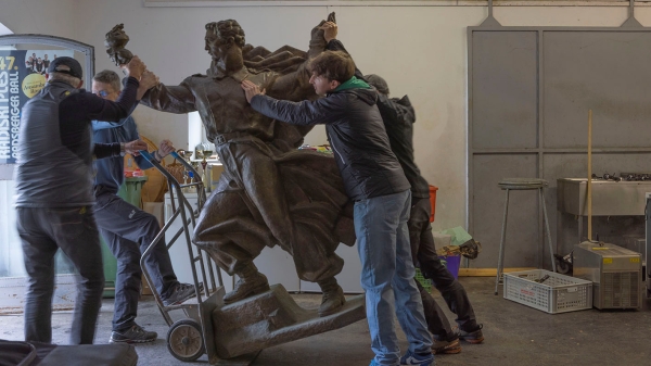 Ein Denkmal einer männlichen Figur wird von einer Gruppe Männer mit Sackkarre transportiert.
