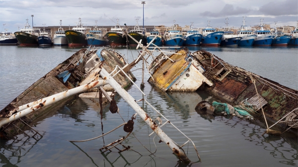 Exhibition poster for “Soil & Water: Mediterranean Crossing”
                                          showing sunken, damaged fishing boats in the foreground and a row of intact boats in a harbor in the background. The photo
                                          originates from the Hotspot Mediterraneo series (2025) by Francesco Bellina. Courtesy of the artist.