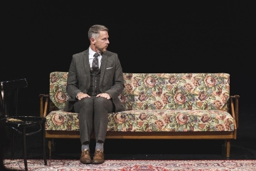 Performer Jim Libby on a sofa on a dark stage